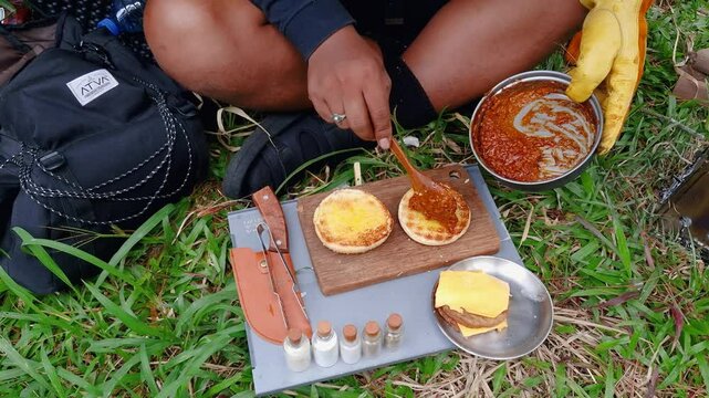 A male adventurer is cooking sauce to make a burger under a flysheet in the dense forest. The concept of bushcraft camping in a dense pine forest for adventure and cooking with simple equipment