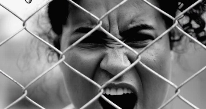 Desperate Young African American woman feeling outrage screaming with rage behind metal fence barrier frowning and open mouth screaming in anger