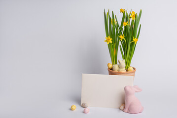 Easter greeting card mockup standing on light background with daffodils in a pot, two bunnies and colorful chocolate eggs creating a spring festive mood