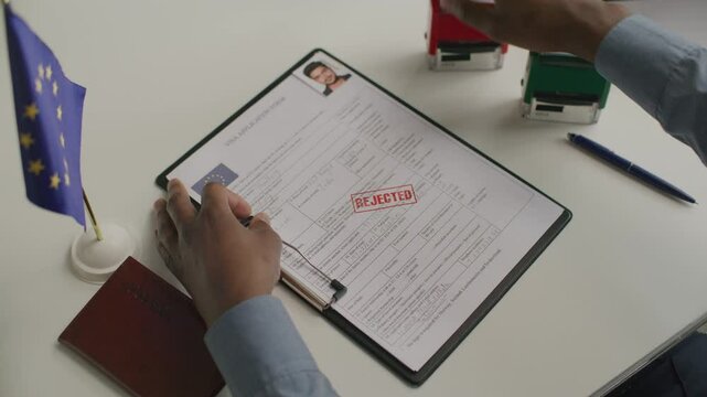 Hands of visa officer stamping red rejection mark on European visa application form on clipboard, with European Union flag and passport on desk beside. Close-up shot