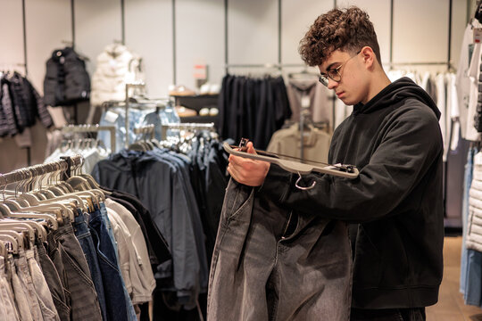 Teenage boy choosing clothes in retail store, young man with glasses and curly hair shopping independently, adolescent exploring fashion options. Copy space