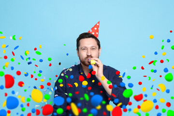 Man in conical paper hat with blower and falling confetti on light blue background. Surprise party