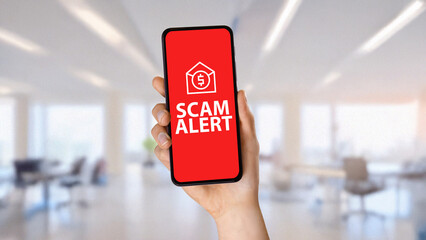 Hand holding smartphone displaying scam alert warning with financial fraud icon on red screen. Concept of online scams, cyber fraud, phishing attacks, identity theft, and digital security awareness.