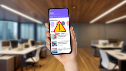Hand holding smartphone displaying scam alert warning over blurred news feed. Concept of cyber fraud, online scam detection, phishing attempts, digital security awareness, and identity theft preventio