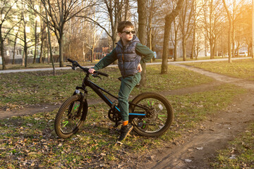Fototapeta premium Child with sunglasses and a bike in a park during golden hour, surrounded by natural scenery. Great for illustrating active kids or outdoor lifestyle moments.