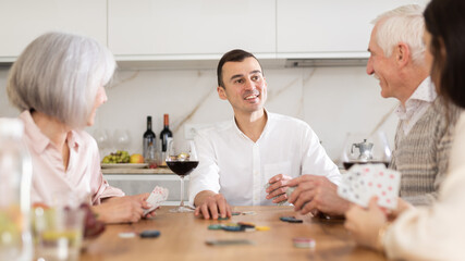 Mature friends is playing card game poker with middle-aged spouses. Married couple pensioners have fun during meeting and gatherings, enthusiastically excitedly fulfill conditions of game with friends