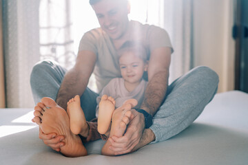 Smiling dad with little girl sitting on bed holding feet with hands