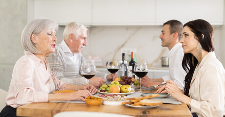 Positive daughter-in-law and mother-in-law exchanging smiles and friendly conversation at dinner table in cozy kitchen, husbands sitting nearby..
