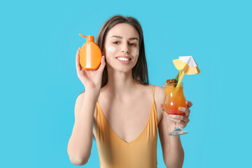 Beautiful young woman with bottle of sunscreen cream and glass of cocktail on blue background