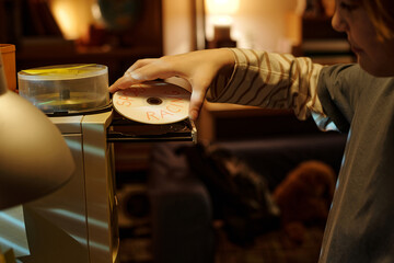 Person inserting a CD into CD player in warm and cozy home setting with soft lighting and home decor elements visible in background