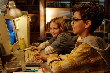 Two children are sitting at a desk, enthusiastically playing a computer game. They are surrounded by various items, illuminated by a desk lamp in a cozy indoor setting