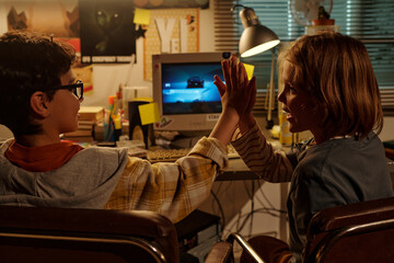 Children sitting at desk high-fiving each other near retro computer screen with dim room lighting creating a nostalgic atmosphere