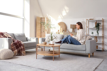 Beautiful young woman reading book on sofa in stylish living room with big window