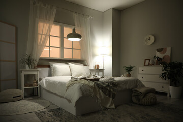 Interior of bedroom with bedside tables, chest of drawers, big bed and glowing lamps in evening