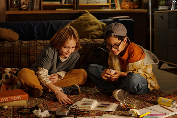 Two children engaging with gadgets while sitting on floor in cozy room, having relaxed expressions...