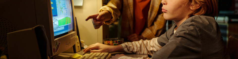 Young child using a computer while receiving direction from an adult in brightly lit room Focused on screen and engaging in educational activities