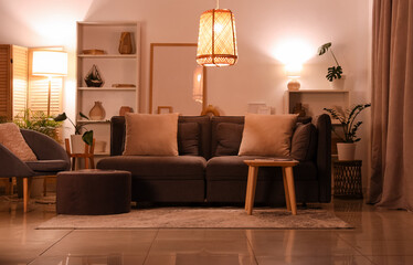 Interior of modern living room with grey sofa, armchair, pouf and glowing lamps at evening