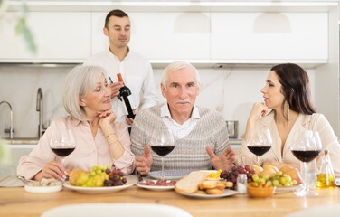 Mature spouses sitting at table, enjoying food and wine, chatting talking laughing with adult son and his girlfriend. In homely atmosphere male and female pensioner celebrate anniversary with guests