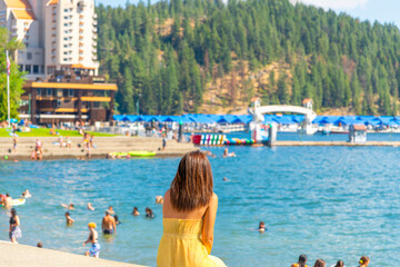 A young woman sits along the boardwalk at city park and beach with the marina and resort in view at summer in Coeur d'Alene, Idaho.