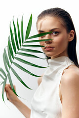 Fototapeta premium A serene young woman with striking features poses with a green palm leaf, showcasing natural beauty and tranquility in a minimalist style.
