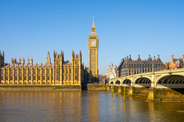 Fototapeta premium Sunlight reflects on the River Thames as the Houses of Parliament and Big Ben stand proudly in London. Westminster Bridge connects the iconic landmarks, depicting a vibrant day.