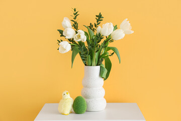 Vase with bouquet of tulip flowers, Easter egg and chick on bedside table near orange wall, closeup