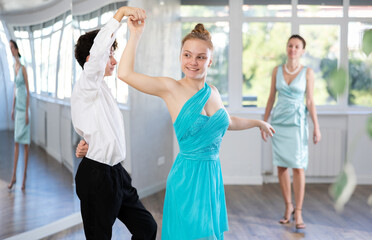 Obraz premium Motivated adolescent ballroom dancers, girl and boy in performance outfit practicing elegant dance moves in pair in bright studio, with female coach supervising in background
