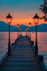 Obraz premium Scenic sunset view of a wooden pier with street lamps, sea, and mountains in the background