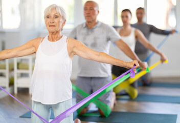 During training, mature woman kneeling performs exercises with colored rubber elastic band. She pulls on and releases expander, elastic band, do tasks to strengthen muscles of arms, shoulders, back