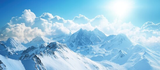 Obraz premium Majestic Snow-Capped Mountains Under a Bright Blue Sky with Fluffy Clouds and Sunlight Illuminating the Scenic Winter Landscape