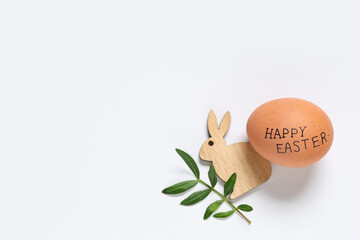Egg with text HAPPY EASTER, wooden bunny and plant leaf on light background