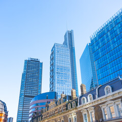 Fototapeta premium Towering skyscrapers dominate the skyline of the City of London financial district. Modern architecture contrasts with historic buildings under a clear blue sky.