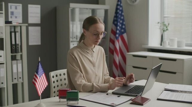 Female visa officer reviewing application form, checking passport, stamping rejection on documents and working on laptop at desk in US embassy