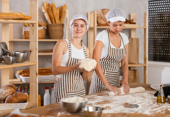 Two skilled female bakers in uniform kneading dough and rolling out dough into sausages before...