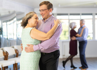 Smiling man supporting mature lady, dancing slow foxtrot in pair during group class in modern...