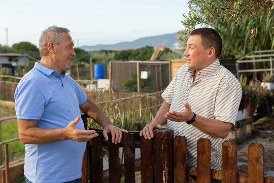 Neighbor conversation. Two smiling men breezily chatting near wooden plank fence of rural house on sunny summer day