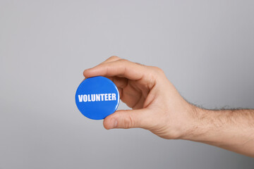 Man holding button badge with word Volunteer on light grey background, closeup