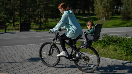 Caucasian woman riding a bicycle with her toddler son sitting behind her in a child seat. 