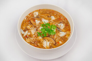 A bowl of hot and sour soup with crab meat, mushrooms, and herbs, garnished with fresh cilantro, served in a white bowl on a matching plate.