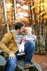 Fototapeta premium Smiling dad looks at a little girl on his knees sitting on a stump in the autumn forest