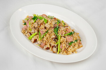 A plate of fried rice mixed with diced ham, leafy greens, and seasonings, served on a white dish,...