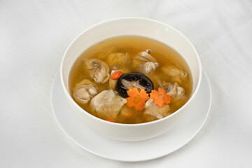 This image shows a white bowl filled with clear chicken soup, containing chicken pieces, a shiitake mushroom, carrot slices, and goji berries in a light broth.