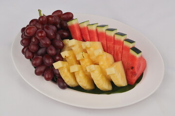 This image shows a white oval plate with an assortment of fresh fruits, including sliced watermelon, pineapple chunks, and a bunch of red grapes, arranged neatly.