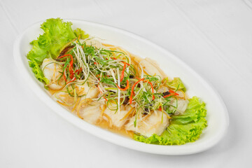 This image shows a white oval plate with steamed fish fillets in a light sauce, garnished with julienned green onions, red chilies, and fresh lettuce leaves.
