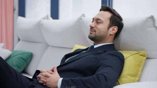 An inspired dreaming man in a formal suit is reclining on a modern white couch with his hands clasped, businessman pensive guy employer relaxing in a positive mood with a satisfactory smile confidence