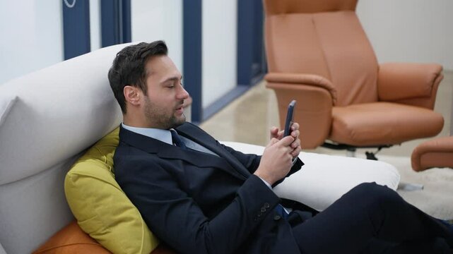 Smiling businessman using a mobile phone, typing an email, gaming on smartphone while relaxing reclines on couch in a positive mood with a satisfactory smile at a luxury executive modern office.