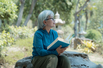 Elderly woman sitting on a rock in a tranquil park, holding a book and gazing off into the...