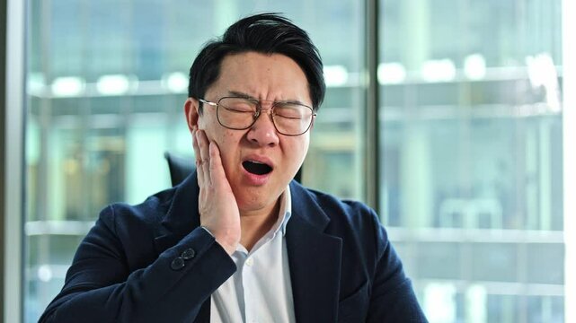 Stressed Korean businessman holds his cheek as he sits in his office. He has tooth pain or ear discomfort.