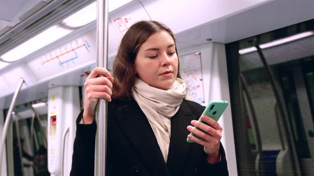 Young Caucasian woman holding a smartphone and standing in a subway train, smiling softly while gripping a pole, wearing a black coat and scarf during an urban commute in public transportation.