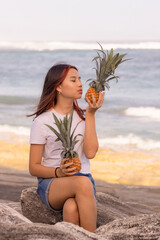 Charming teenager girl sitting on the stone, holding two pineapples. Kissing lips. Closed eyes. Organic fruit concept.